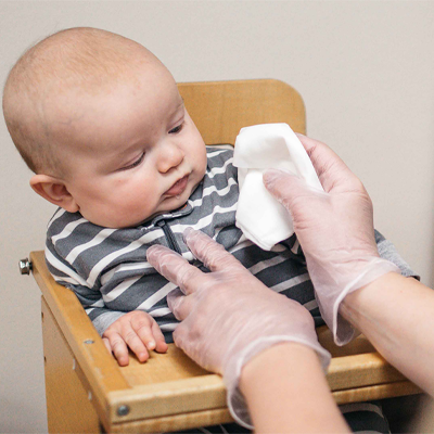 A baby sitting in a high chair looks at a tissue held by an adult wearing AMMEX Clear Vinyl Exam Latex Free Disposable Gloves (Case of 1000).