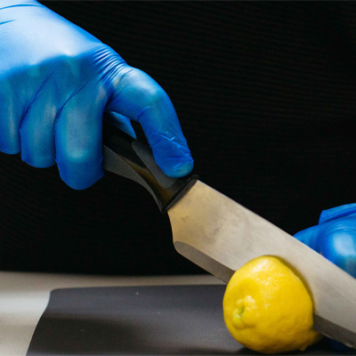 A person wearing Gloveworks Blue Vinyl Industrial Latex Free Disposable Gloves (Case of 1000) cuts a lemon on a black cutting board with a stainless steel knife.