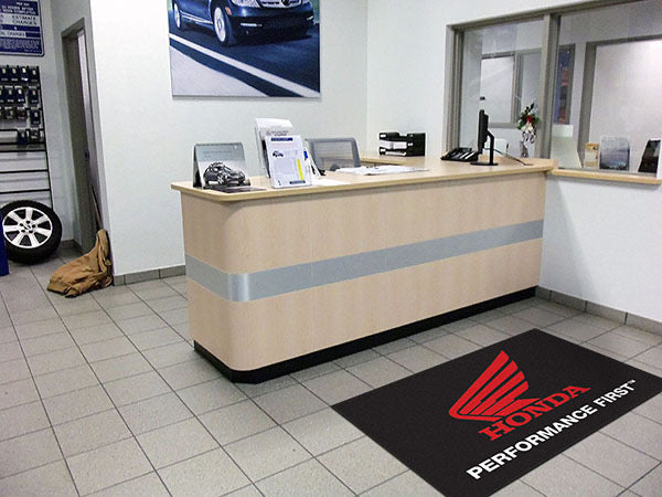 A reception area in a Honda dealership with a desk, papers, a phone, and a large Custom Carpet Logo Mat featuring the Honda logo.