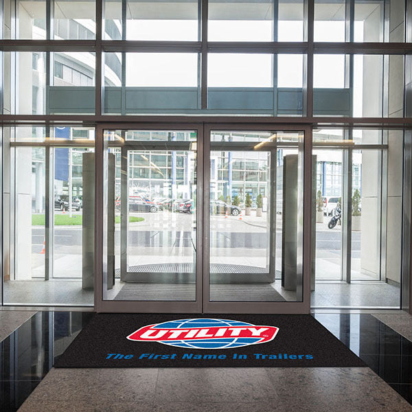 Glass entrance doors of a modern building with a "Custom Carpet Logo Mat" on the floor, showcasing the building’s access to a street with parked cars visible outside.