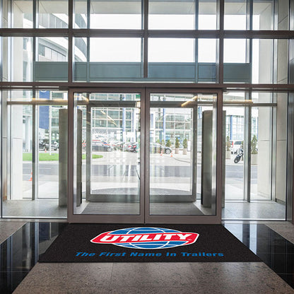 Glass entrance doors of a modern building with a "Custom Carpet Logo Mat" on the floor, showcasing the building’s access to a street with parked cars visible outside.