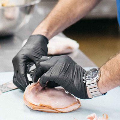 A person in X3 Black Nitrile PF Ind Gloves trims chicken breasts on a steel countertop, wearing a wristwatch.