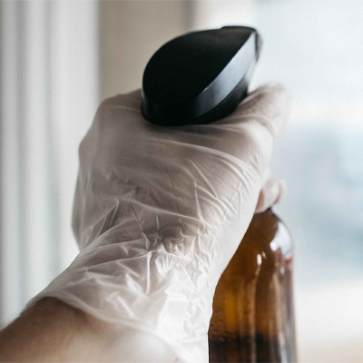 A hand wearing a Gloveworks Clear Vinyl Industrial Latex Free Disposable glove holding a brown glass bottle with a black cap near a window.