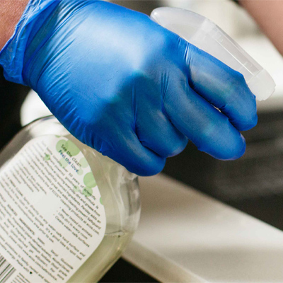 A hand in Gloveworks Blue Vinyl Industrial Latex Free Disposable Gloves holds a plastic container with a green label, examining its contents.