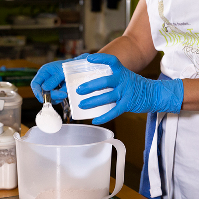 Person wearing X3 Ultra Blue Nitrile PF Ind Gloves scoops powder from a container into a large clear mixing bowl on a table.