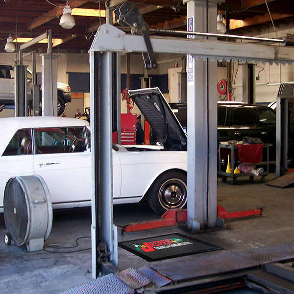 A mechanic works on a white classic car at a car service station, with open bays and scattered Hi Res Logo Scraper Mats visible in the background.