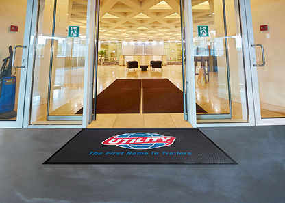 View from an entrance with open glass doors leading into a lobby with a geometric patterned ceiling and a Hi Res Logo Scraper Mat branded "utility.