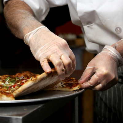 A chef wearing X3 Stretch Hybrid Poly Disposable Gloves slices a freshly baked pizza on a metal tray in a dimly lit kitchen.