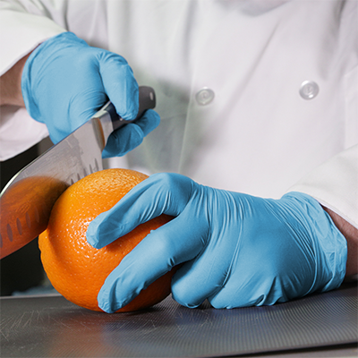 A person in a lab coat and AMMEX Stretch Synthetic Blue Vinyl PF Exam Gloves cutting an orange with a knife on a dark surface.