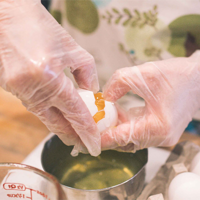 Hands in Gloveworks Clear Vinyl Industrial Latex Free Disposable Gloves cracking an egg over a metal bowl, with other cooking ingredients and tools nearby.