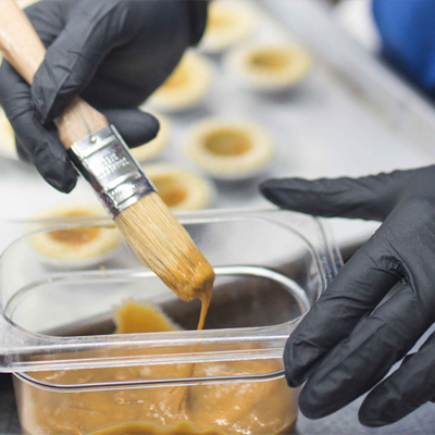 A person wearing X3 Black Nitrile PF Ind Gloves uses a brush to apply a brown glaze to pastries on a tray in a professional kitchen.