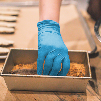 A person wearing X3 Blue Nitrile Industrial Latex Free Disposable Gloves presses a mixture into a rectangular baking dish.