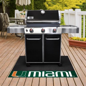 A black gas grill with three knobs and a University of Miami Hurricanes Grill Mat reading "umami" on a patio with green foliage and outdoor furnishings in the background.