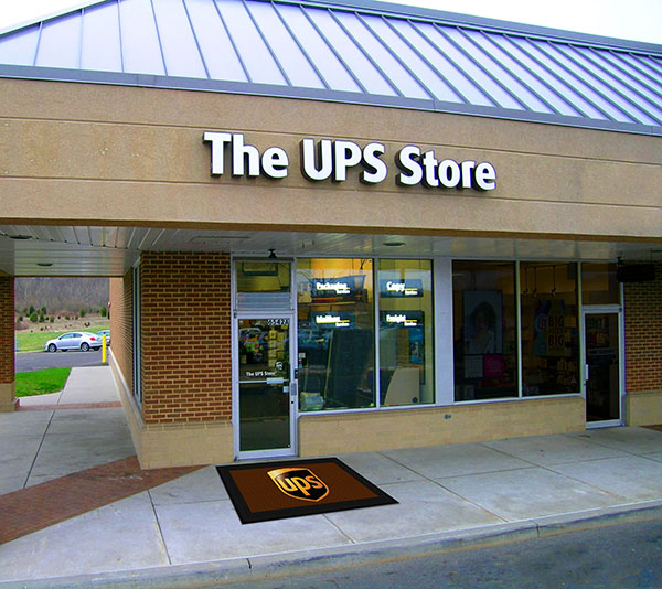 Exterior view of a UPS store with visible signage, entry door, and a Hi Res Logo Scraper Mat in front of the store.