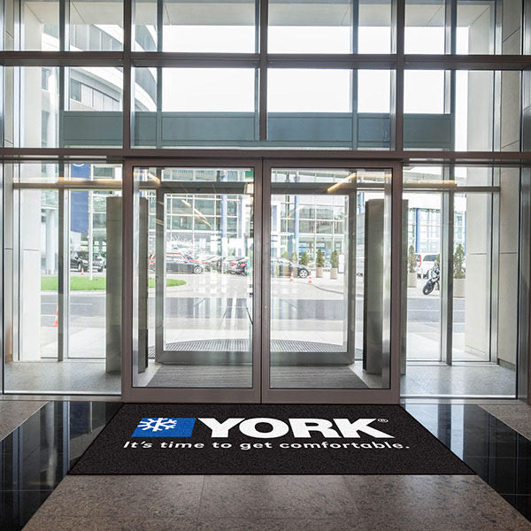 Glass entrance doors of a modern building with the "york" logo and slogan on Custom Carpet Logo Mats, visible street and vehicles outside.