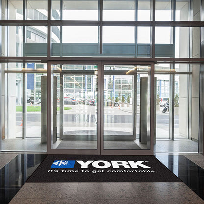 Glass entrance doors of a modern building with the "york" logo and slogan on Custom Carpet Logo Mats, visible street and vehicles outside.