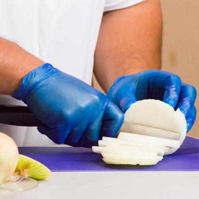 A person in Gloveworks Blue Vinyl Industrial Latex Free Disposable Gloves is slicing an onion on a purple cutting board.