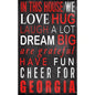 Decorative sign with phrases "in this house we love, hug, laugh a lot, dream big, are grateful, have fun, cheer for georgia" in white/red letters on a black background.