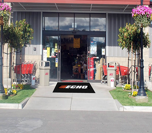 Entrance of a hardware store with an Hi Res Logo Scraper Mats welcome mat, shopping carts to the left, potted plants, and visible interior shelves stocked with items.