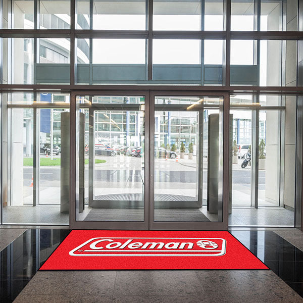 View from inside a modern building looking out through glass doors, featuring a red Custom Carpet Logo Mat in the entrance.