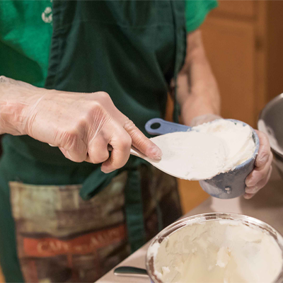 Person in an apron scooping flour from a bowl with a measuring cup while wearing AMMEX Clear Vinyl Exam Latex Free Disposable Gloves (Case of 1000) in a kitchen.