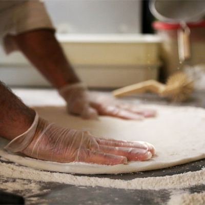 A person wearing X3 Stretch Hybrid Poly Disposable Gloves flattens pizza dough on a floured surface in a kitchen.