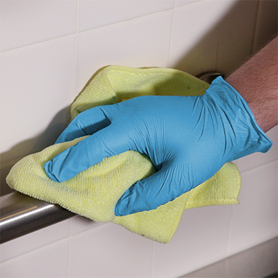 A person wearing AMMEX Stretch Synthetic Blue Vinyl PF Exam Gloves cleans a tile surface with a yellow cloth.