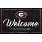 A decorative welcome sign featuring a prominent letter "g" logo, styled with a distressed black wood background and the words "welcome to our home" in white cursive and block letters.