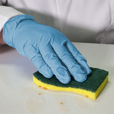 A person wearing AMMEX Stretch Synthetic Blue Vinyl PF Exam Gloves (Case of 1000) cleans a surface with a yellow and green sponge.