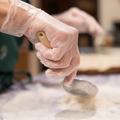 Hands in AMMEX Clear Vinyl Exam Latex Free Disposable Gloves shaping dough with a spoon and flour on a kitchen counter.