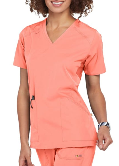 Woman in MULTI-POCKET V-NECK TOP C12106 scrubs smiling, standing with one hand slightly raised, visible waist up.