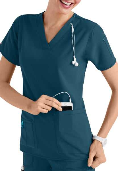Nurse in CROSS FLEX V-NECK MEDIA SCRUB TOP C12110 using smartphone with stethoscope around neck, smiling and looking to her side.