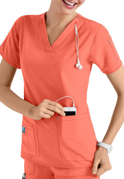 Smiling female healthcare worker in Coral Cross Flex V-Neck Media Scrub Top C12110 using a smartphone, with a stethoscope around her neck, isolated on a white background.