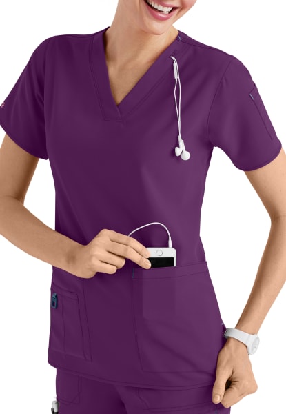 A smiling healthcare worker in a CROSS FLEX V-NECK MEDIA SCRUB TOP C12110, placing a device with headphones into her pocket.