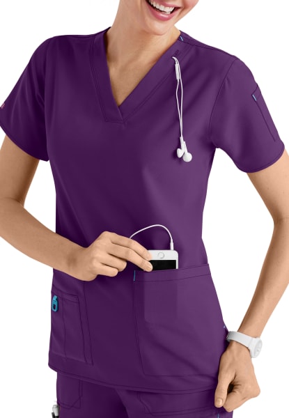 Smiling female healthcare worker in CROSS FLEX V-NECK MEDIA SCRUB TOP C12110 placing a smartphone in her pocket, wearing a stethoscope around her neck.