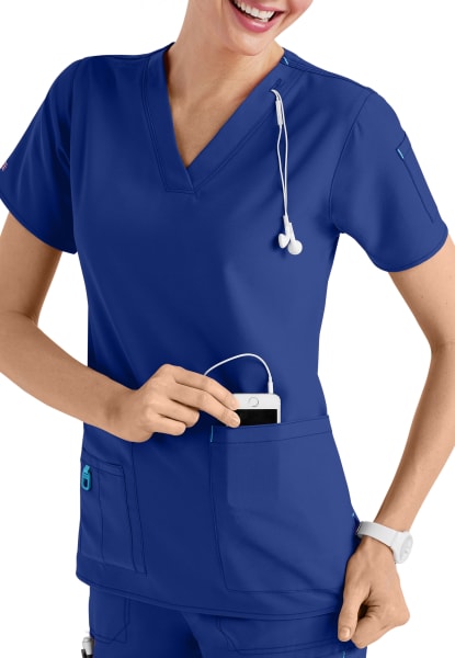 A smiling healthcare worker in a CROSS FLEX V-NECK MEDIA SCRUB TOP C12110 with a stethoscope around her neck, inserting a smartphone into her pocket.