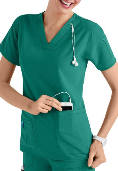 A smiling healthcare professional in a CROSS FLEX V-NECK MEDIA SCRUB TOP C12110 puts a device in her pocket, wearing a stethoscope around her neck.