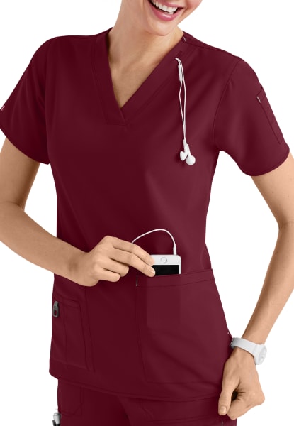 A smiling healthcare worker in a CROSS FLEX V-NECK MEDIA SCRUB TOP C12110 uniform using a mobile device with headphones attached.