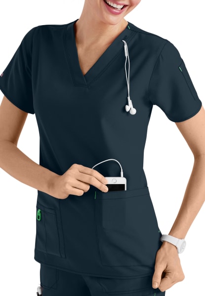A smiling female nurse in a CROSS FLEX V-NECK MEDIA SCRUB TOP C12110, placing a device into her pocket, with a stethoscope around her neck.