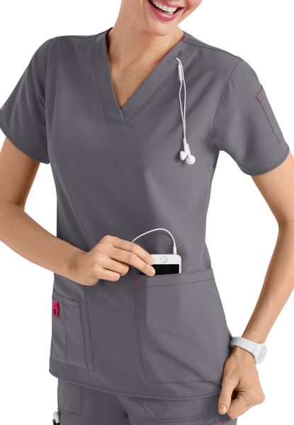 A smiling healthcare professional in a CROSS FLEX V-NECK MEDIA SCRUB TOP C12110 puts a mobile device into her pocket, with a stethoscope around her neck.