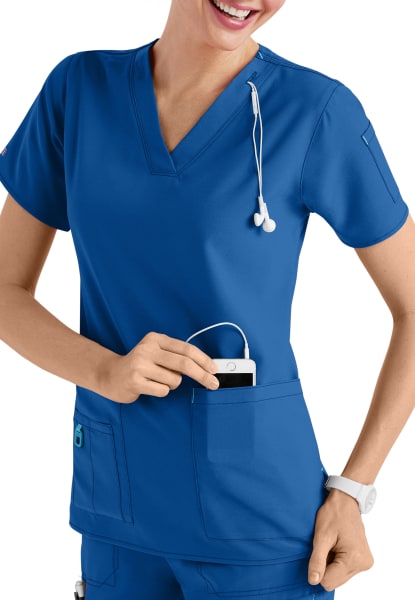A smiling healthcare worker in a CROSS FLEX V-NECK MEDIA SCRUB TOP C12110 places a digital device into her pocket, with a stethoscope around her neck.