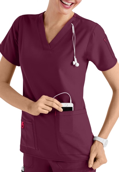 A smiling female healthcare professional in a CROSS FLEX V-NECK MEDIA SCRUB TOP C12110, inserting a device into her pocket, with a stethoscope around her neck.