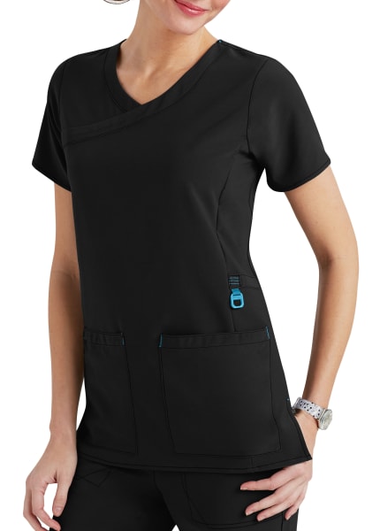 A woman wearing a black medical scrub top with blue accents, and a wristwatch, standing with hands partially in pockets.