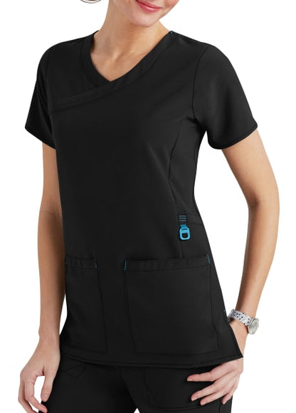 A woman wearing a black medical scrub top with blue accents, and a wristwatch, standing with hands partially in pockets.