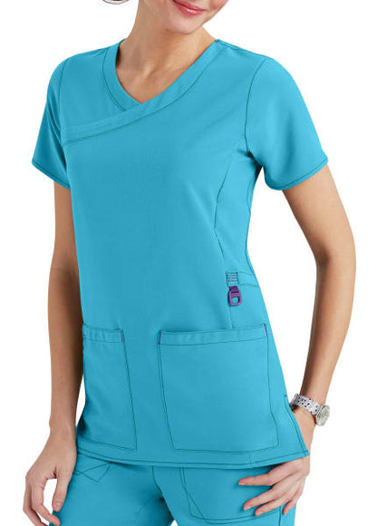 A woman in teal scrubs with a v-neck top featuring large pockets, standing against a plain background.