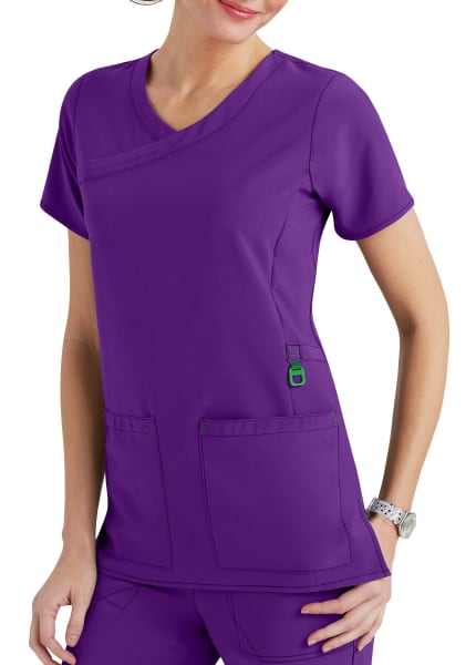 Woman in purple medical scrubs with a v-neck, short sleeves, and multiple pockets, standing with one hand lightly resting on her hip.